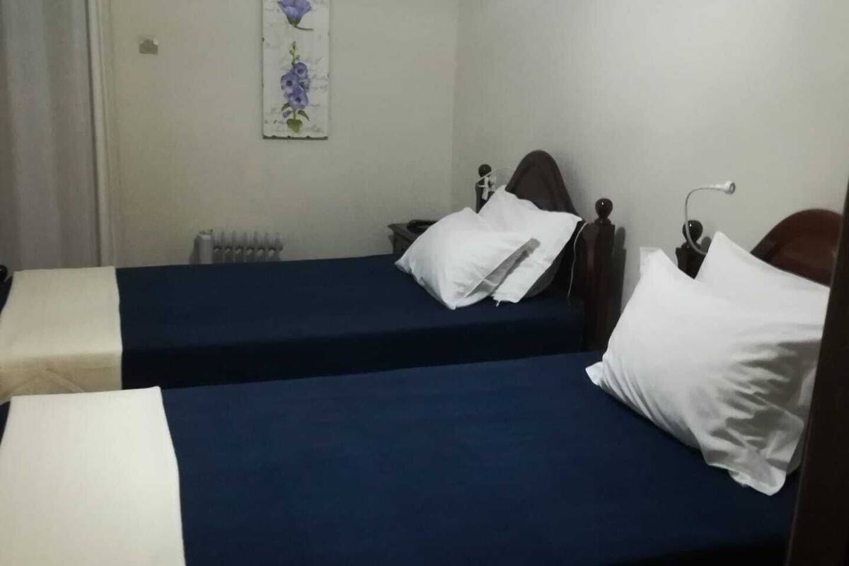 A room with two beds, each with a blue mattress and white pillows. There is a radiator on the wall and a small table with a lamp.