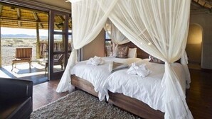 A bedroom with a four-poster bed, white linens, and a view of the beach through large glass doors. - Hoodia Desert Lodge (Sesriem)