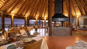 A spacious dining area with a high thatched ceiling, stone walls, and a fireplace. Tables are set for dining with candles and tableware. - Hoodia Desert Lodge (Sesriem)
