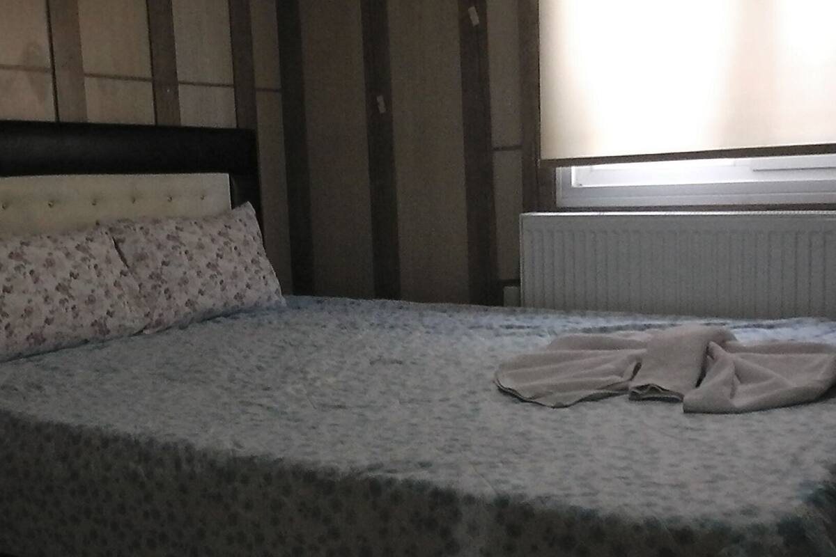 Una cama con un cojín estampado con motivos florales y una colcha, un radiador y una ventana con persianas.