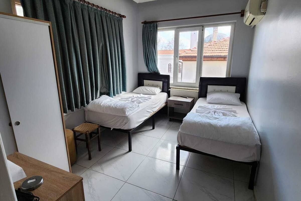 Habitación de hotel con dos camas individuales, un escritorio de madera, una silla pequeña, una ventana con cortinas y una lámpara en el techo.