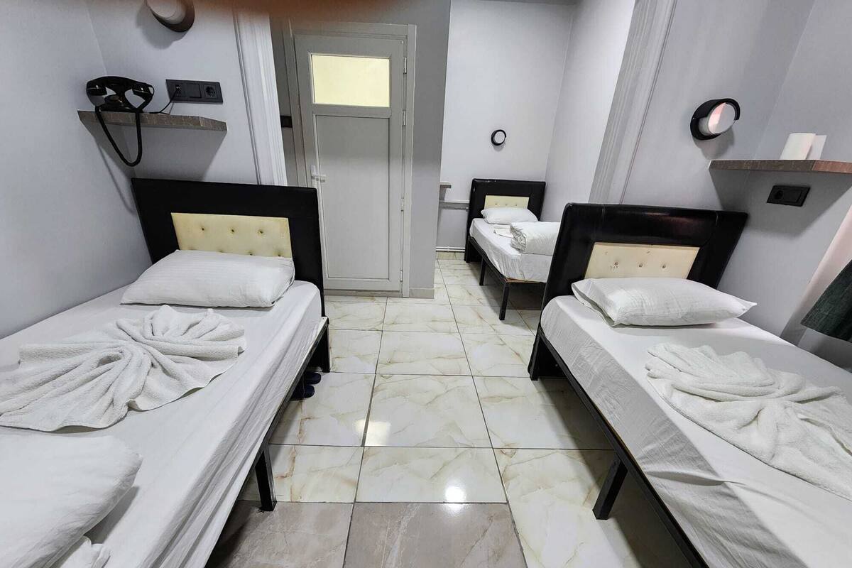 Habitación de hotel con dos camas individuales, una puerta, una mesita pequeña y un estante fijado a la pared.