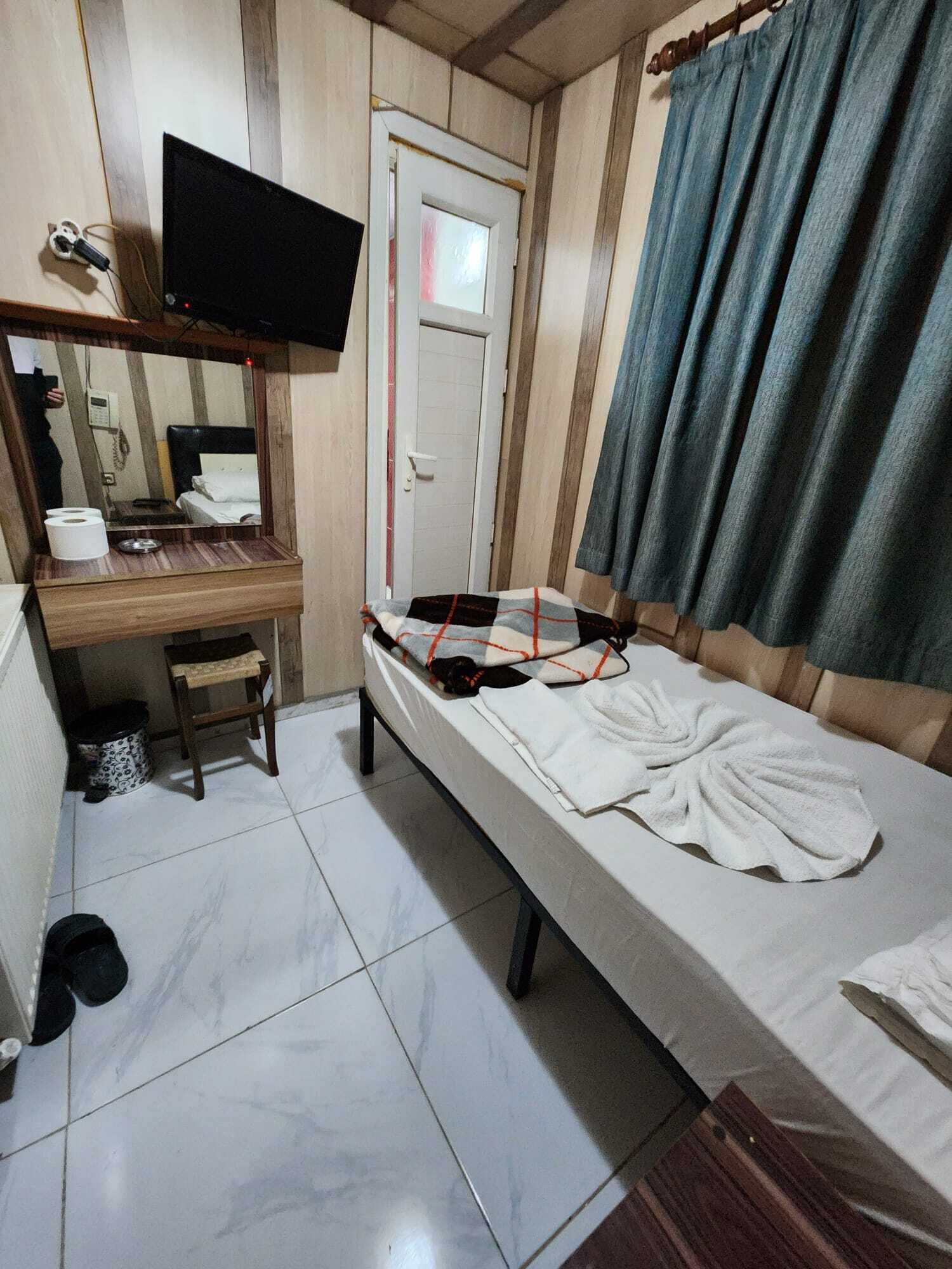 Quarto individual com uma cama, uma escrivaninha, televisão fixada na parede, uma porta e uma cortina.