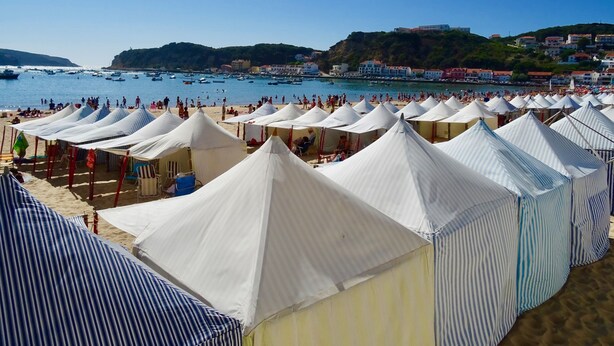 Ein Strand mit vielen weißen Zelten und gestreiften Sonnenschirmen, einem klaren blauen Himmel und Booten im Wasser.