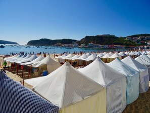 A beach with numerous white tents and striped umbrellas, a clear blue sky, and boats in the water.
