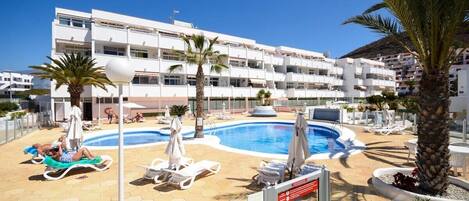 A hotel with a swimming pool, sun loungers, and palm trees.