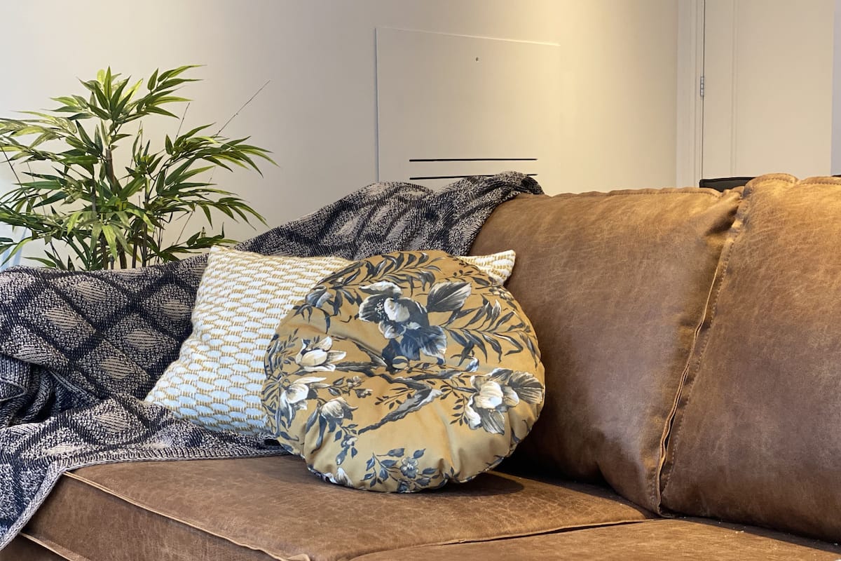 A brown leather sofa with floral and patterned cushions, a woven throw blanket, and a potted plant.