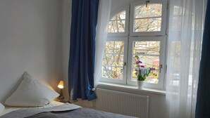 Ein Schlafzimmer mit Bett, Kissen, einer Nachttischlampe, einem Fenster mit Vorhängen und Blick auf Bäume und Blumen.
