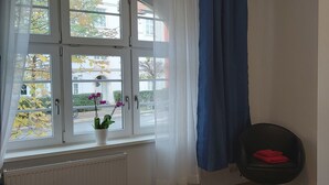 Ein Zimmer mit großem Fenster, Heizkörper, Topfpflanze, schwarzem Stuhl und Schrank.