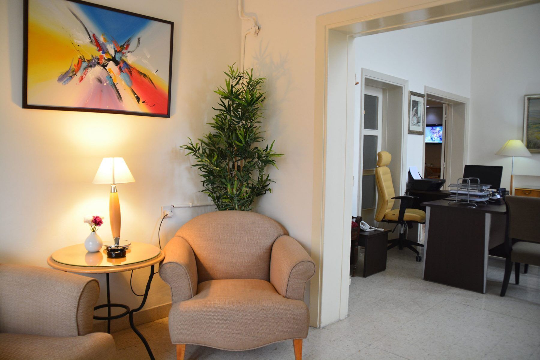 A hotel room with a beige armchair, a small round table with a lamp, a potted plant, and a desk with a chair and computer.