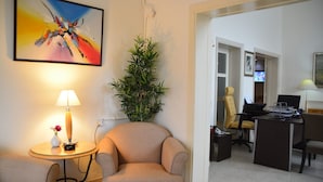A hotel room with a beige armchair, a small round table with a lamp, a potted plant, and a desk with a chair and computer. - The Y Hotel Amman (Amman)