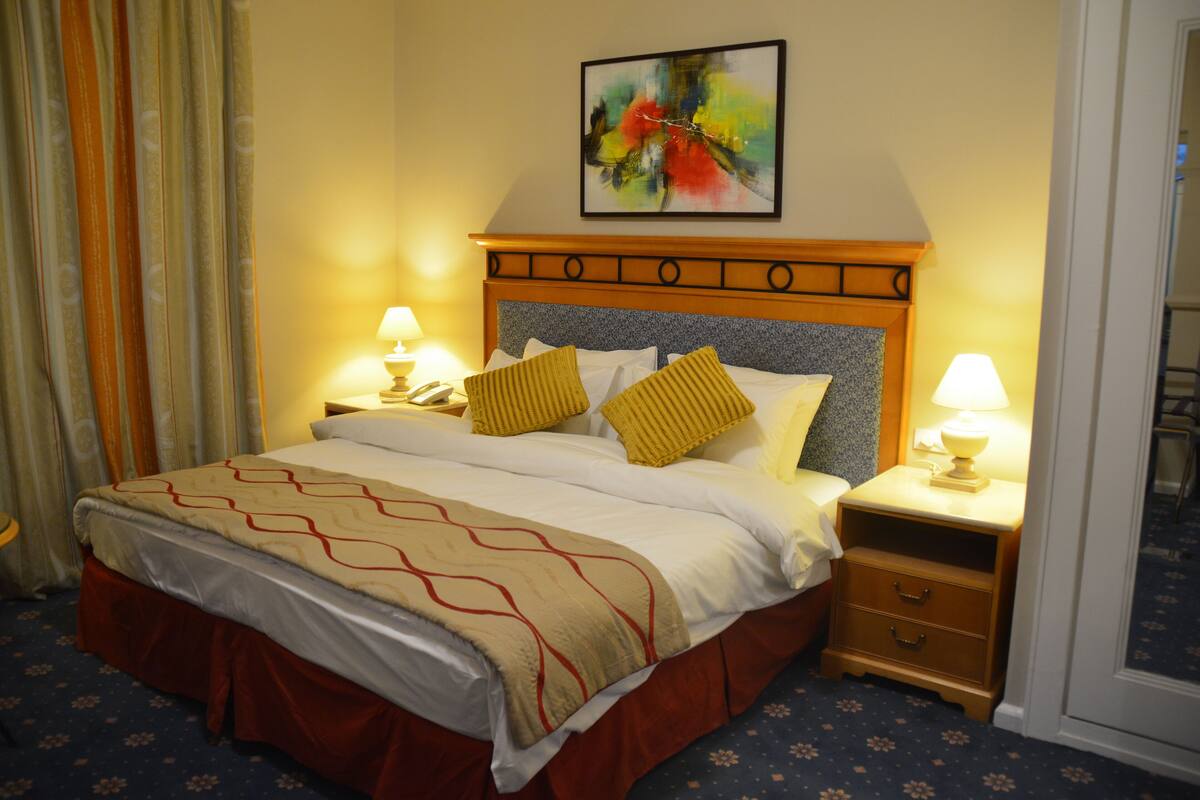 Une chambre d’hôtel avec un lit, deux tables de chevet, une commode, un tableau au mur et une fenêtre avec des rideaux.