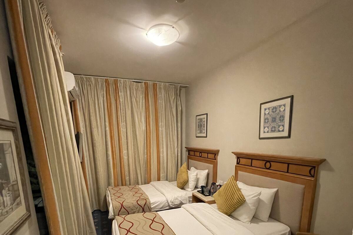 Une chambre d’hôtel avec deux lits, une tête de lit en bois, une petite table, une chaise, un luminaire au plafond et une fenêtre avec des rideaux.