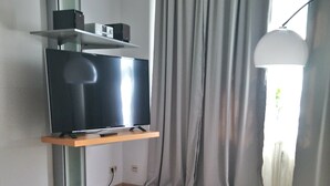 A modern living room with a flat-screen TV on a wooden shelf, grey curtains, and a white pendant light.