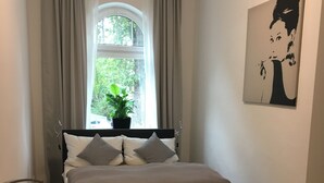 A bedroom with a bed, a window with curtains, a potted plant, and a wall-mounted artwork.
