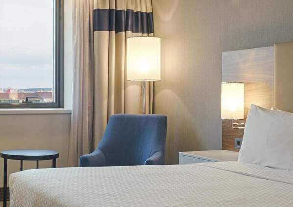 A hotel room with a bed, a chair, a small table, a lamp, and a view of the city through the window.