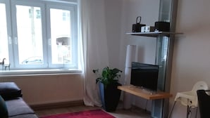 A living room with a sofa, a red rug, a TV, a plant, and a window with curtains. - Fewo Reißhausstraße 23 - 2 Schlafzimmer (Erfurt)