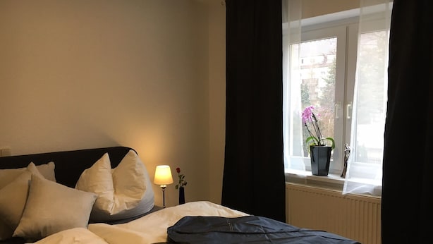 A neatly made bed with pillows, a bedside lamp, and a window with curtains. - Fewo Reißhausstraße 23 - 2 Schlafzimmer (Erfurt)