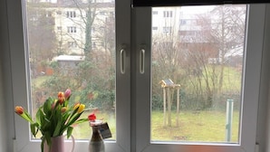 A window with a view of a garden, a vase of tulips, a bottle, and a plate with fruits. - Fewo Reißhausstraße 23 - 2 Schlafzimmer (Erfurt)