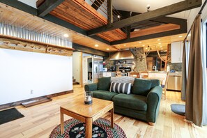 Un salon chaleureux avec un canapé vert, une table basse en bois et un tapis à motifs. On aperçoit une cuisine à l’arrière-plan, équipée d’appareils électroménagers modernes.