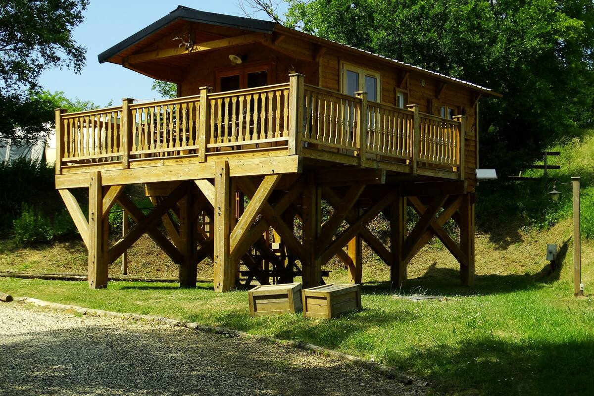 A wooden cabin with a balcony, built on stilts, surrounded by grass and trees.
