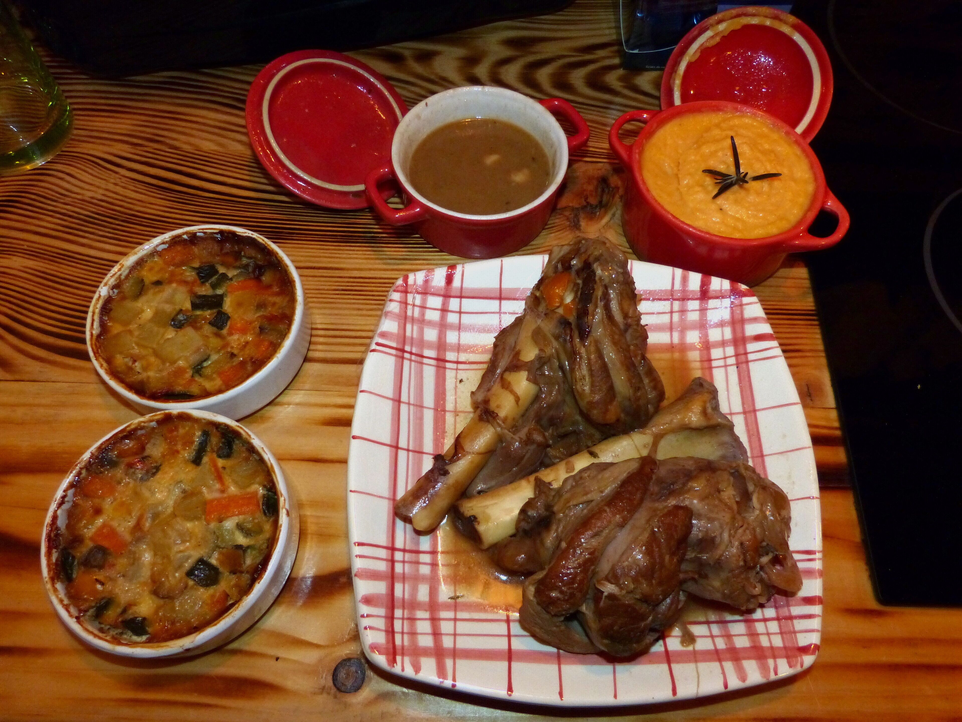 A wooden table with various dishes including a plate of roasted lamb, two bowls of soup, and a red pot with a creamy dish.