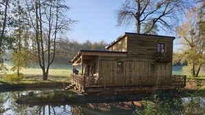 Une cabane en bois dotée d’un balcon, construite sur pilotis au-dessus d’une étendue d’eau, et entourée d’arbres et de verdure.
