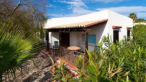 Un edificio blanco con tejado de tejas rojas, un patio con mesa y sillas, y un pequeño jardín con palmeras.