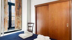 A room with a bed, two rolled towels, a wooden wardrobe, a chair, and a window with a view of buildings. - National Theater House 24 (Porto)