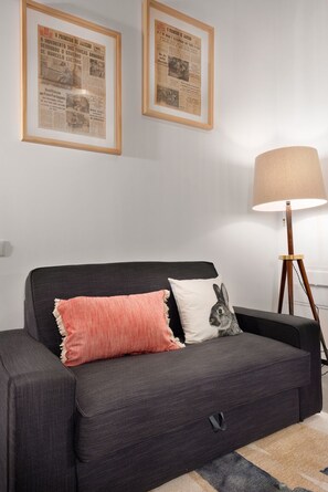 A modern living room with a dark grey sofa, two framed historical newspapers, a lamp, and a rabbit-themed pillow.