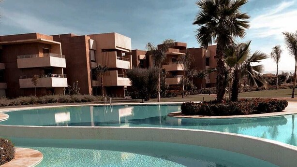 Une piscine à l’eau claire, entourée d’un espace paysager agrémenté de palmiers et, au loin, d’un ensemble de bâtiments.