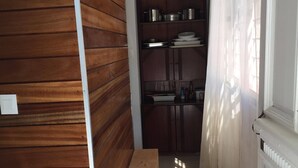 A narrow hallway with wooden paneling on one side, a bench, and a kitchen area with cabinets and a window with sheer curtains. - Nossa Casa Vossa Casa (Tarrafal)