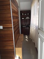 Un pasillo estrecho con paneles de madera en una pared, un banco y una zona de cocina con armarios y un ventanal con cortinas traslúcidas.