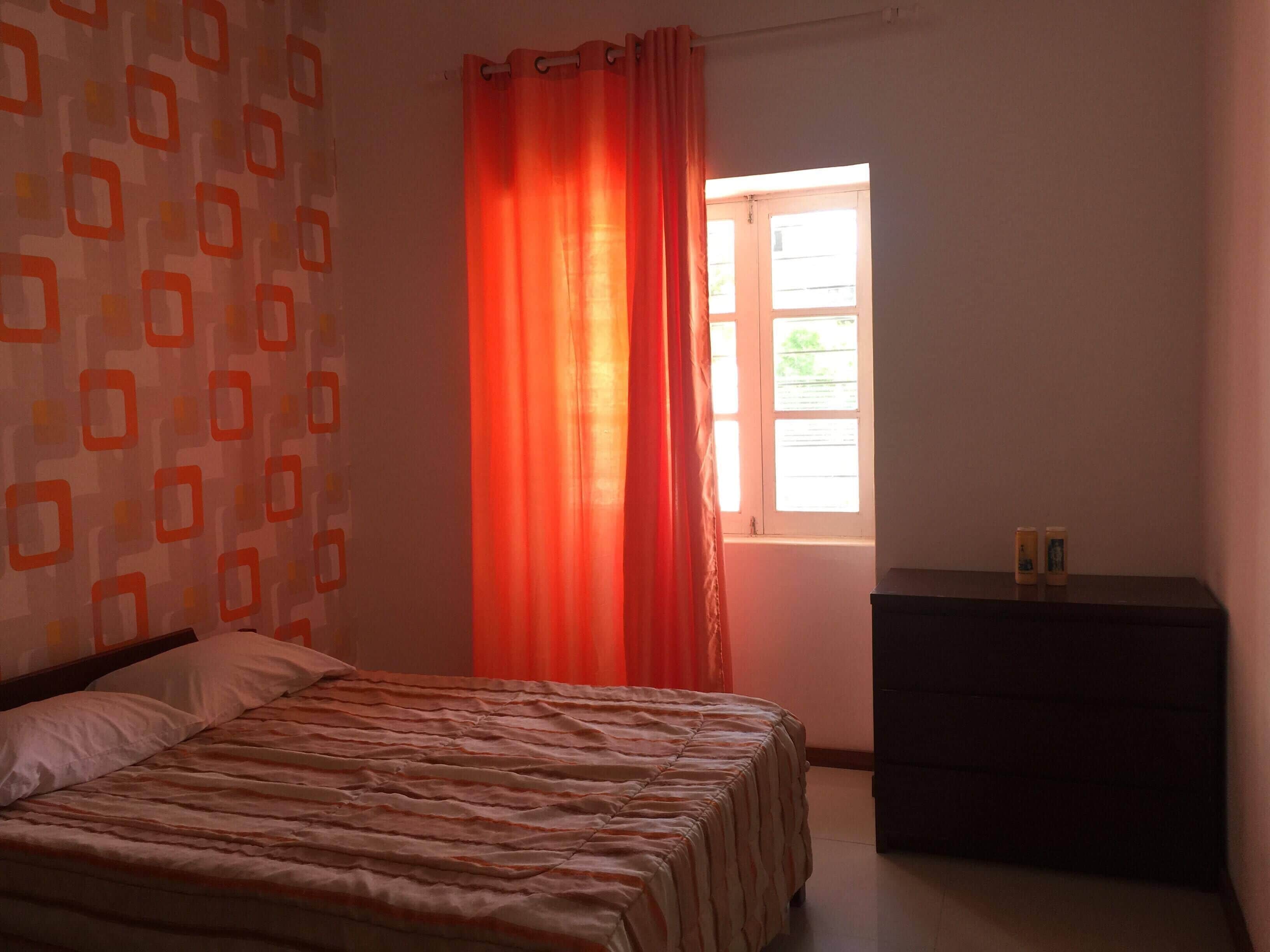 A bedroom with a bed, a window with curtains, a dresser, and a patterned wall.