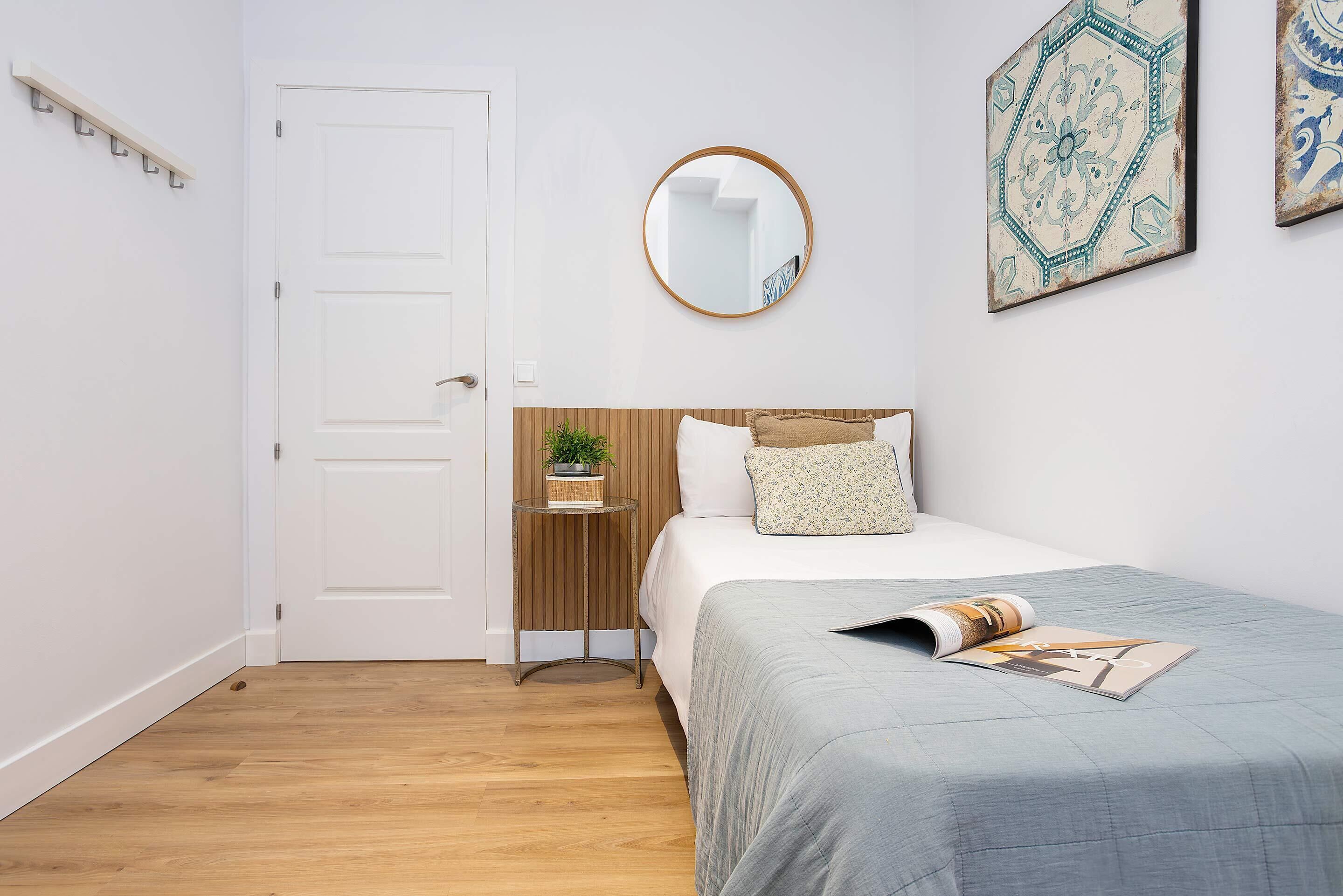 A bedroom with a bed, a bedside table with a plant, a round mirror, and a wall with decorative tiles.