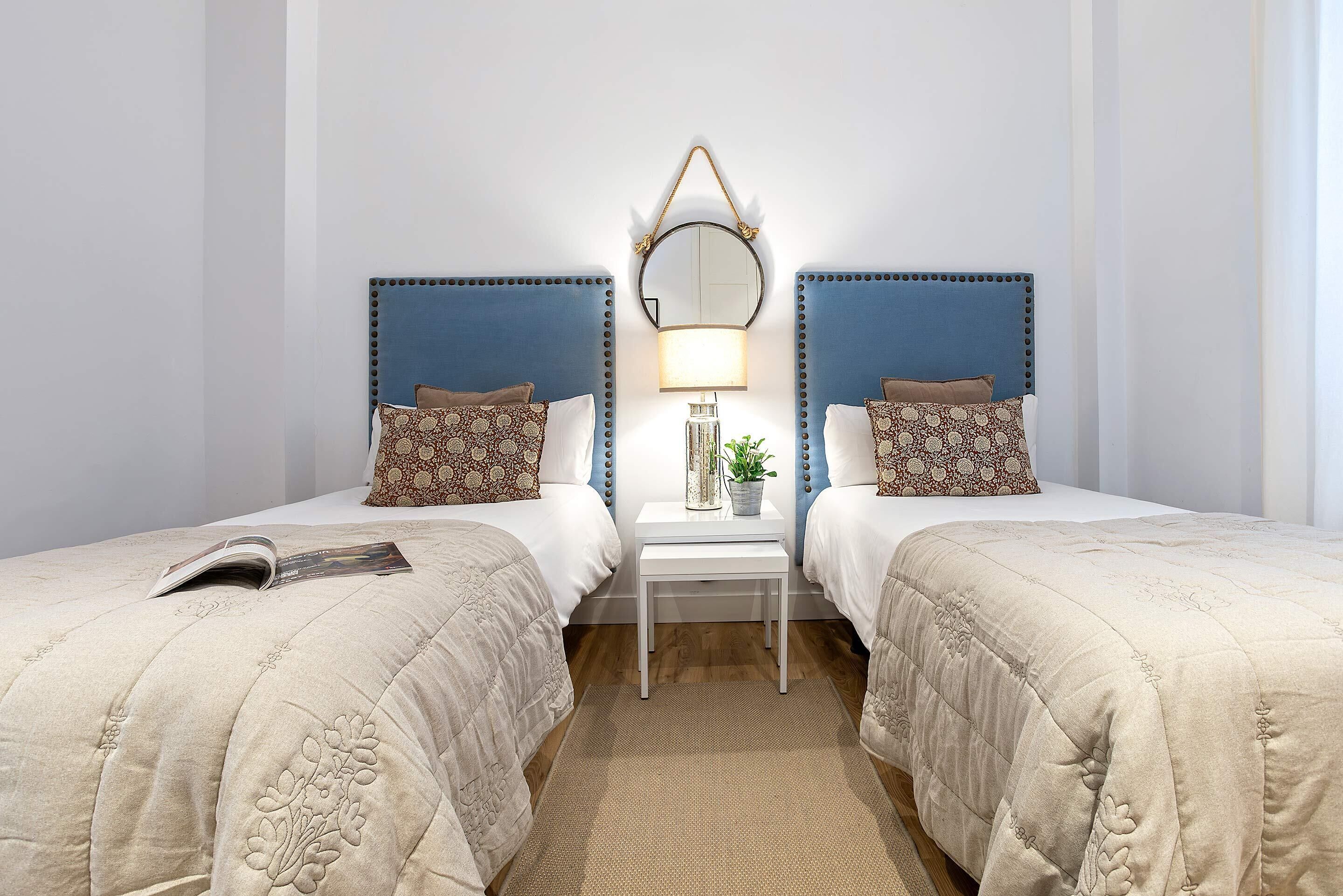 A hotel room with two beds, a bedside table with a lamp, and a mirror.