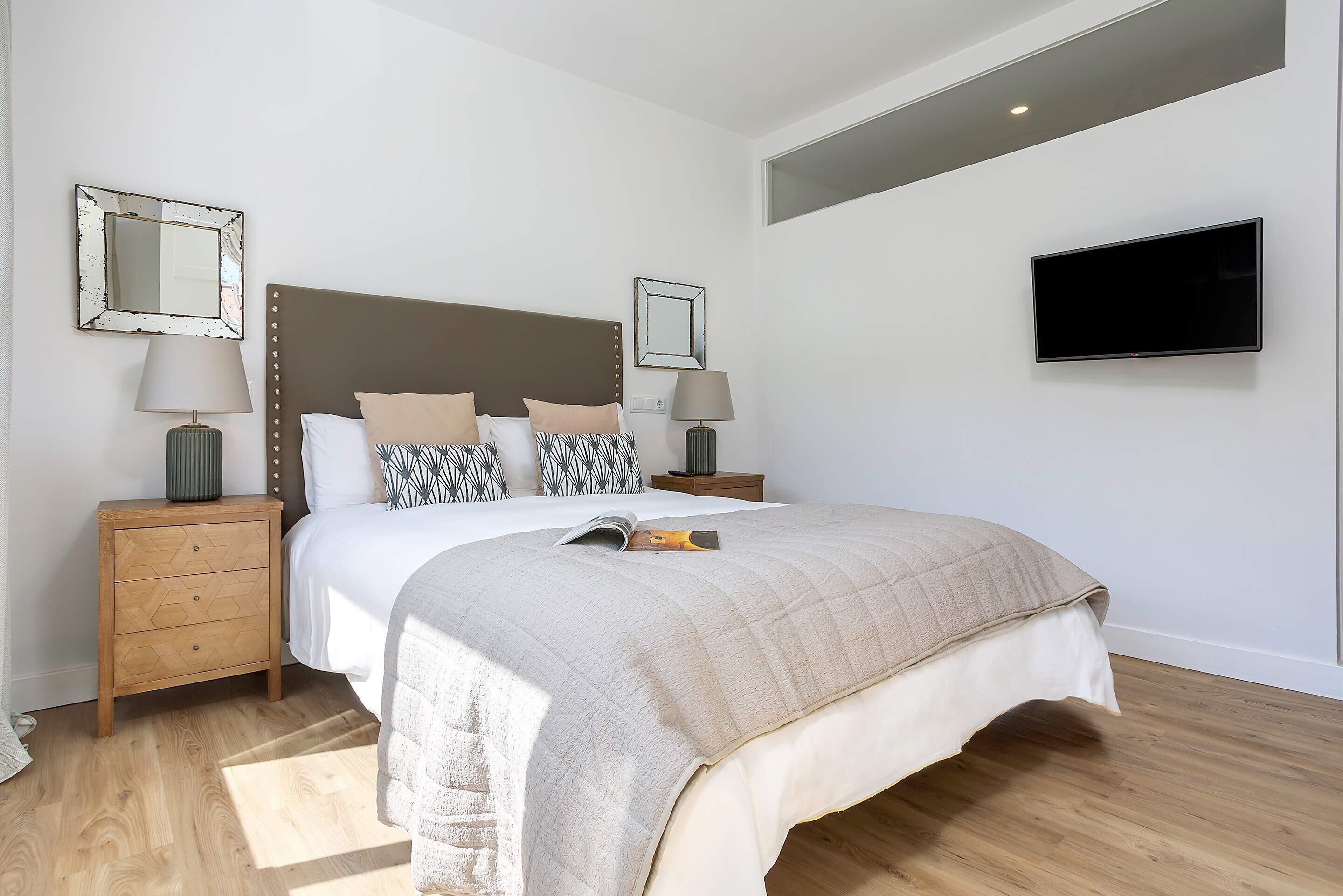 A modern bedroom with a large bed, two bedside tables with lamps, a flat-screen TV, and a large mirror on the wall.