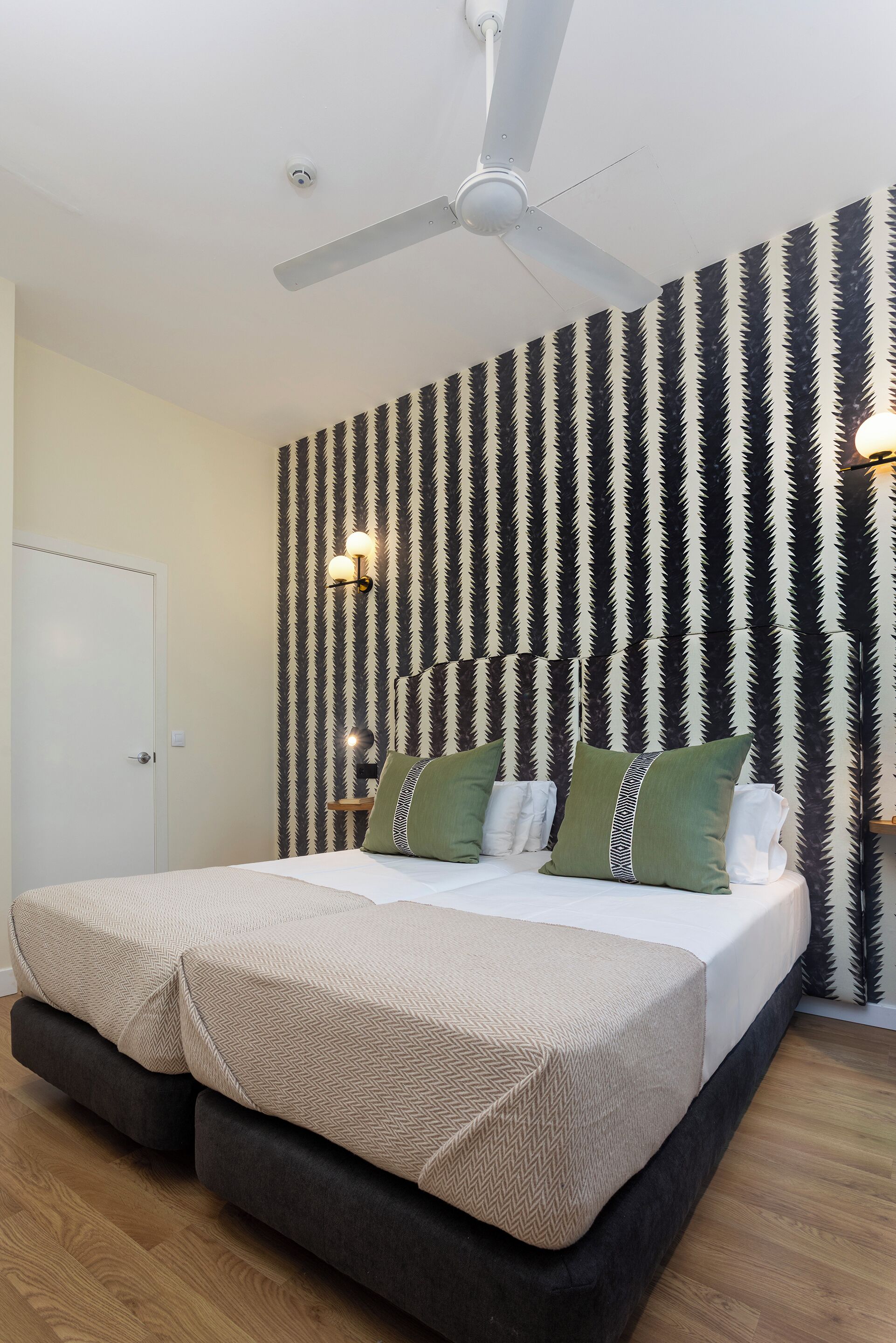 Uma cama de casal com duas almofadas, um ventilador de teto e uma parede com padrão listrado.