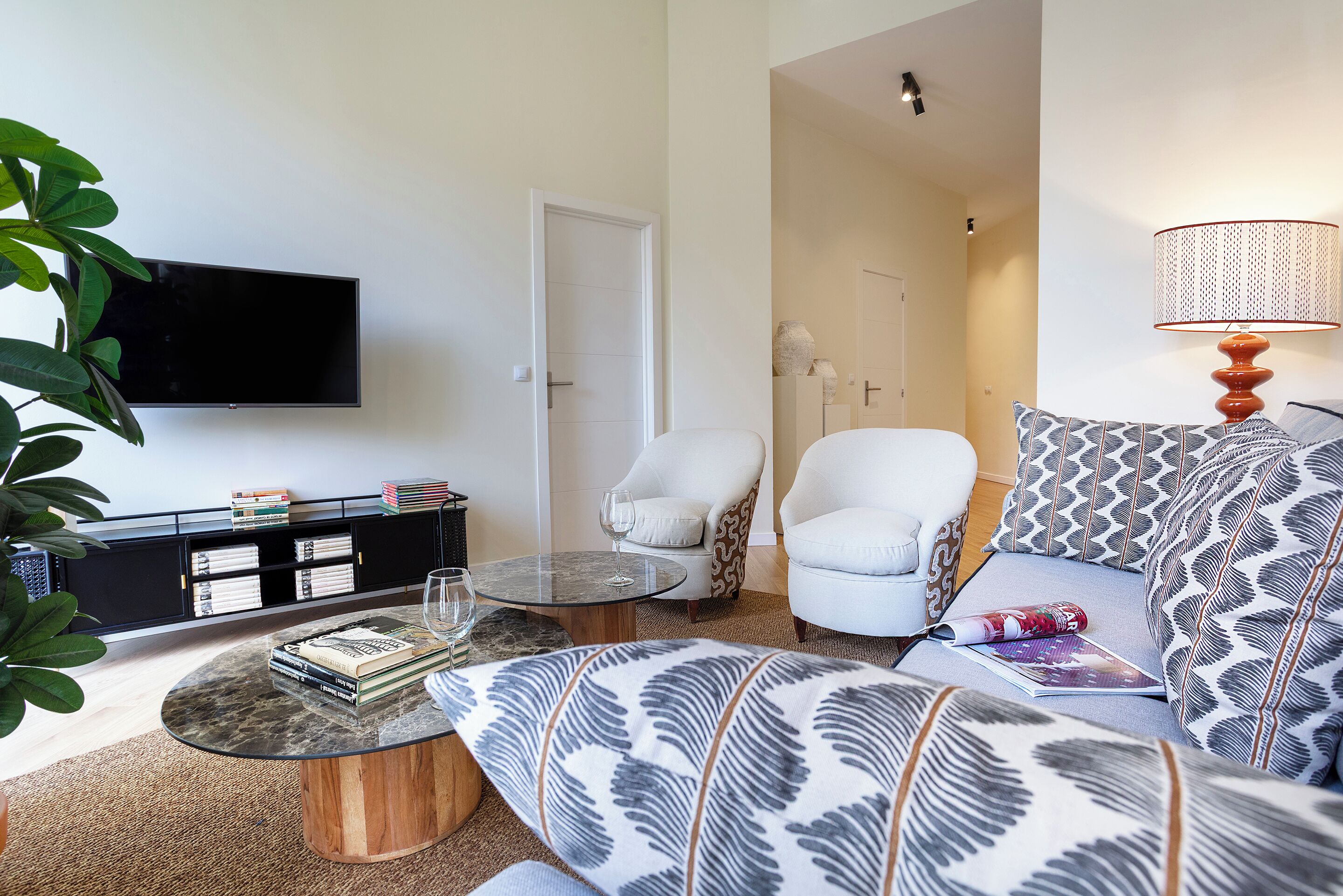 Una sala moderna con un televisor de pantalla plana, una mesa de centro, un sofá con cojines estampados, un sillón blanco y una estantería.