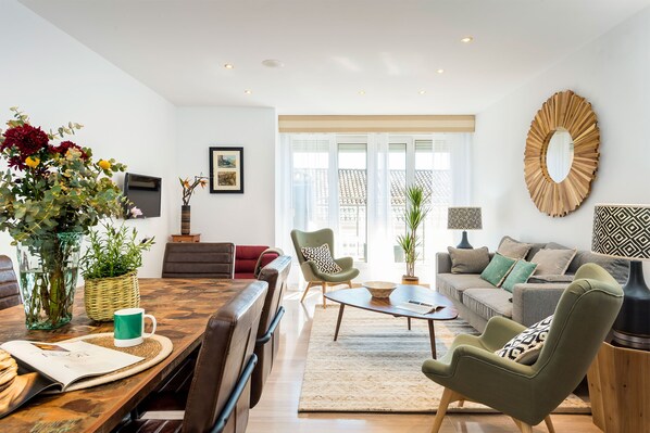 Een moderne woonkamer met een houten eettafel, een bank en een salontafel. Aan de muur hangt een grote spiegel en het raam biedt uitzicht op gebouwen.