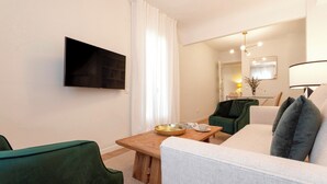 A modern living room with a flat-screen TV mounted on the wall, a wooden coffee table, a green armchair, a white sofa with grey cushions, and a dining area with a small table and chairs. - Genteel Home Velarde (Seville)