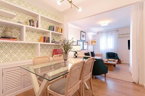 Uma sala de estar moderna com uma mesa de jantar de vidro, cadeiras de vime, uma estante, uma poltrona verde e um abajur.