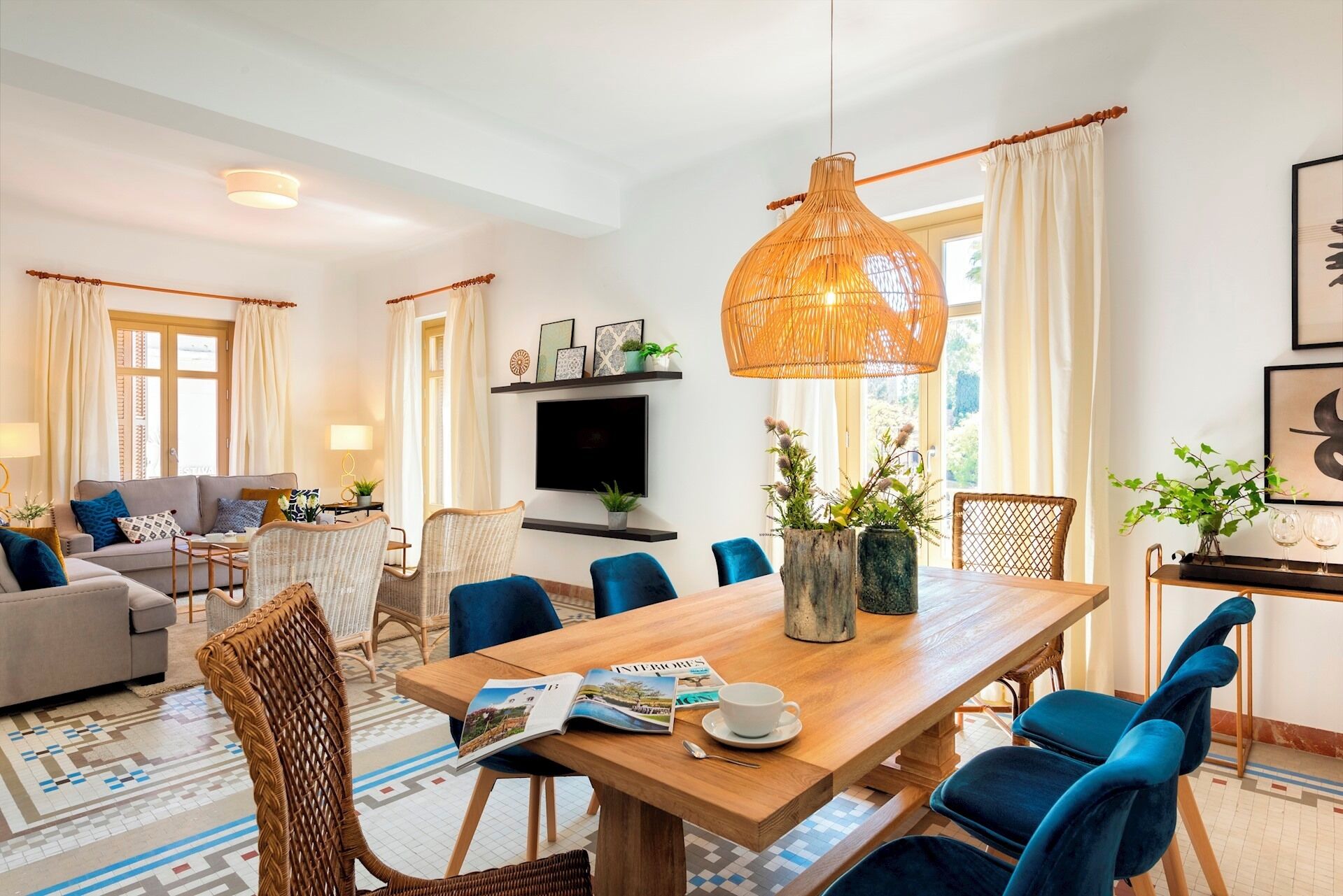 Uma sala de estar moderna com área de jantar, destacando uma grande mesa de madeira, cadeiras azuis, uma cadeira de vime e um pendente de rattan.