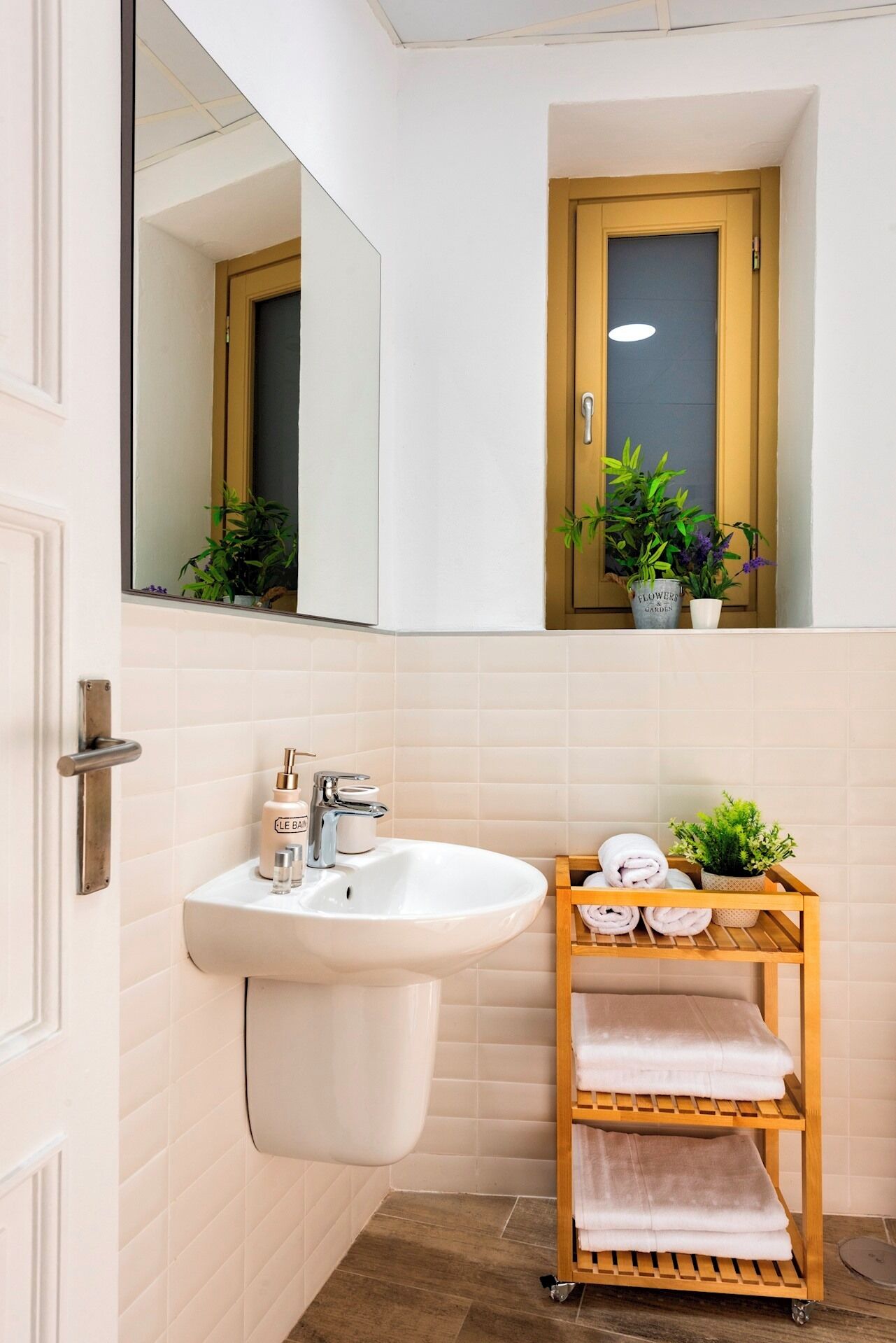 Une salle de bain avec un lavabo blanc, un miroir, une étagère en bois avec des serviettes et une plante en pot.