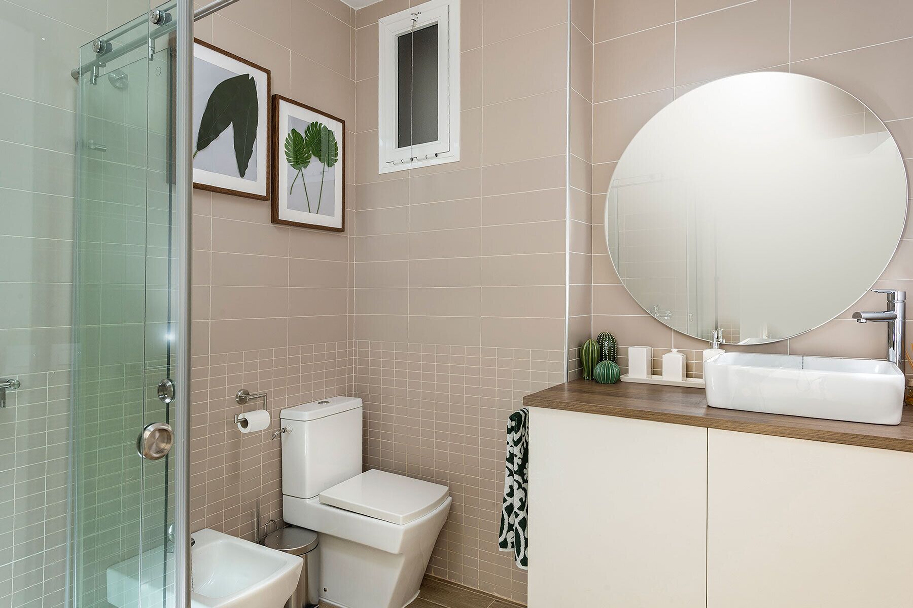 A bathroom with a glass shower enclosure, a toilet, a sink with a mirror, and framed plant pictures on the wall.