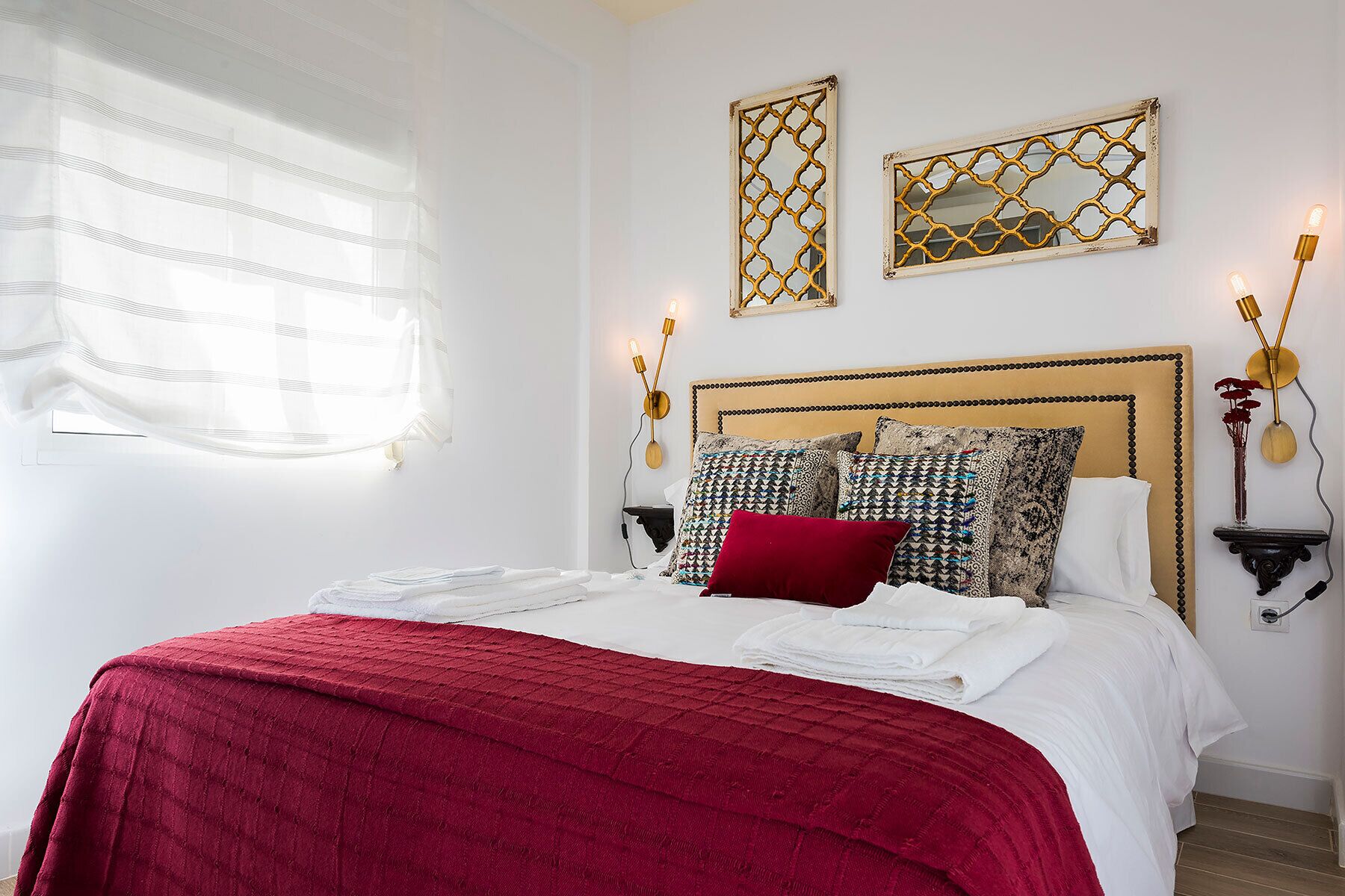 A neatly made bed with a red blanket, two wall-mounted lamps, and two decorative mirrors above it.