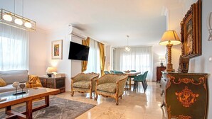 Un salon spacieux comprenant un canapé, des fauteuils, une table basse et un téléviseur. On y trouve également un coin repas avec une table et des chaises, ainsi qu’un meuble décoratif avec une lampe.