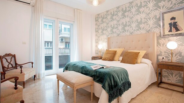 Une chambre à coucher comprenant un lit, des tables de chevet, un banc, une chaise, une fenêtre avec des rideaux et un mur orné d’un motif floral.