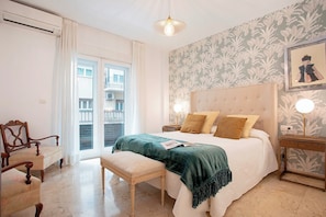 Ett sovrum med en säng, sängbord, en bänk, en stol, ett fönster med gardiner och en vägg med ett blommönster.
