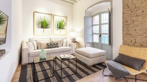A living room with a sofa, ottoman, coffee table, and a wicker chair. There are two framed wall art pieces, a lamp, and a window with a view.